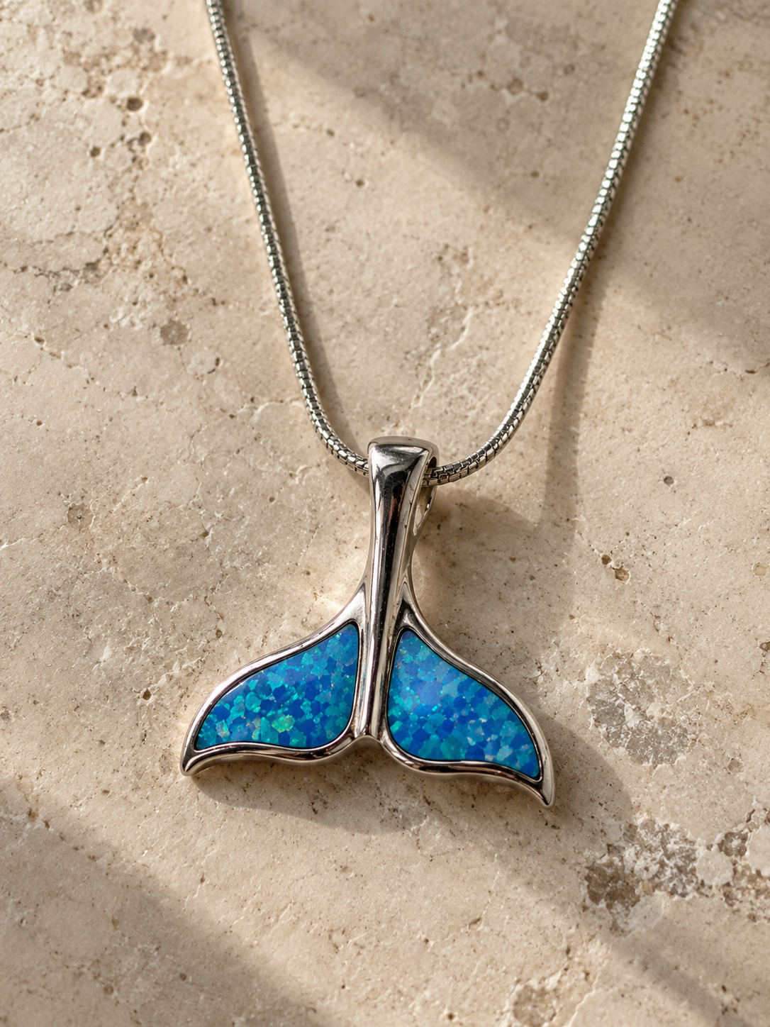 Sterling silver whale tail pendant necklace with vibrant blue opal inlay, photographed in natural light on a neutral stone surface