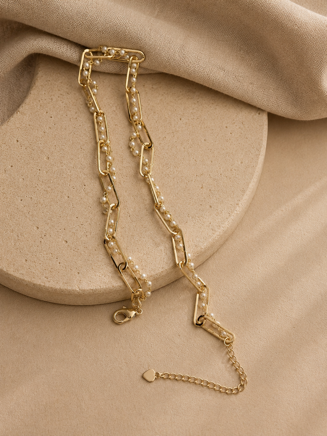Gold paperclip chain choker necklace with delicate pearl accents displayed on a beige stone tray and soft linen background. Elegant minimalist jewelry flat lay in warm neutral tones by Mancerat.