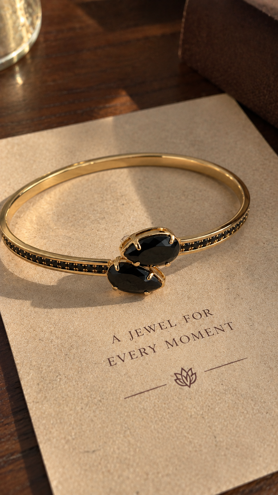 A gold-toned bangle bracelet with two black oval stones and small black stones along the band, displayed on a beige card over a dark wooden surface.