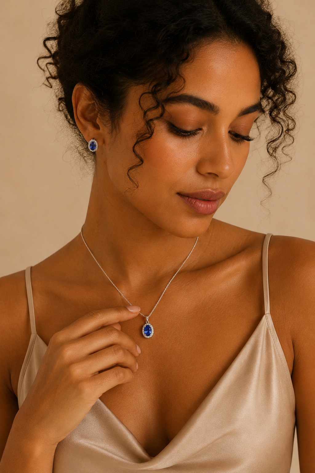 Elegant tanned woman with curly hair wearing a sterling silver blue oval halo pendant necklace and matching stud earrings while gently touching the necklace against a warm beige studio background.