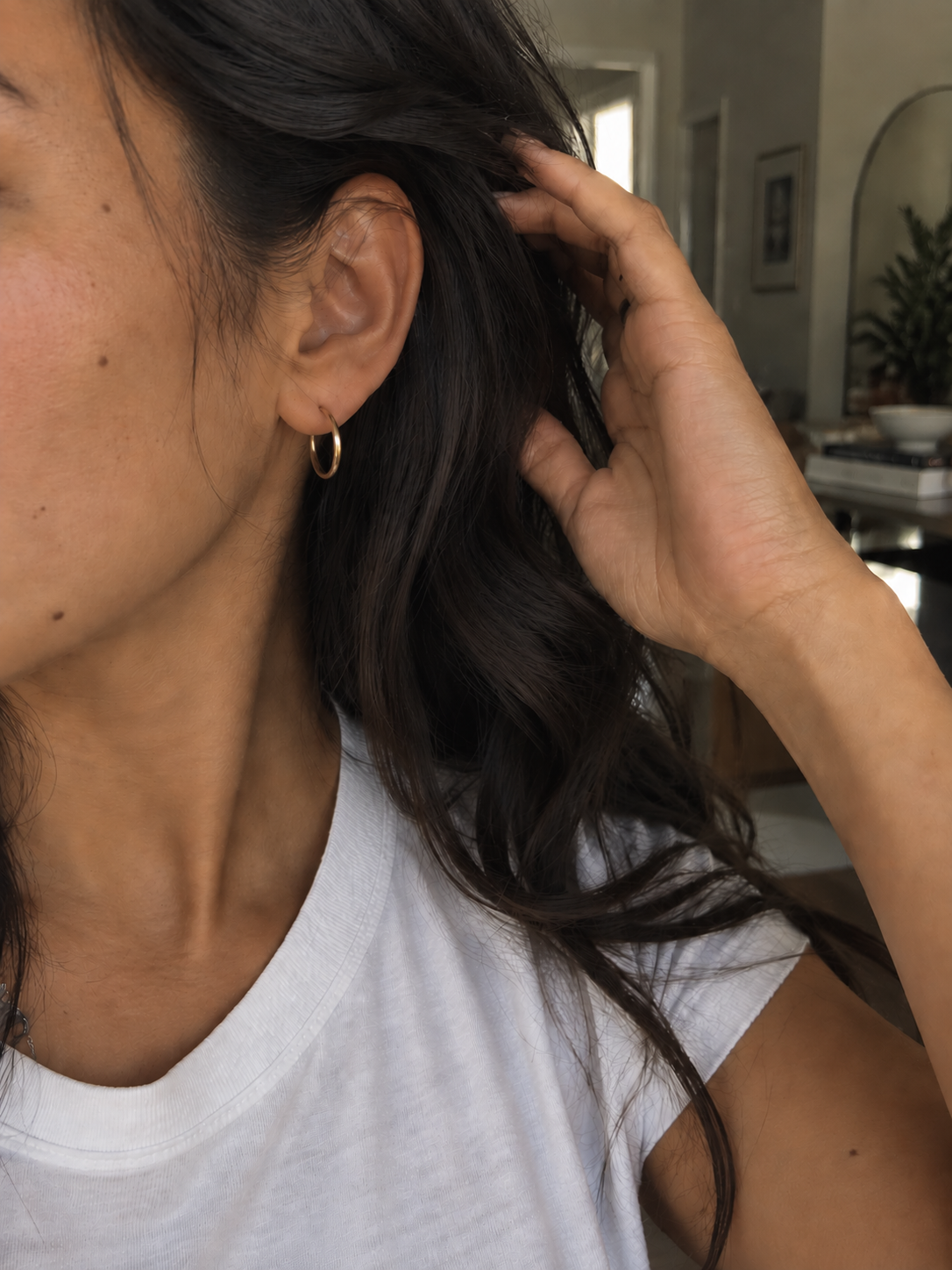 Woman wearing small gold hoop earrings with a white T-shirt in natural window light.