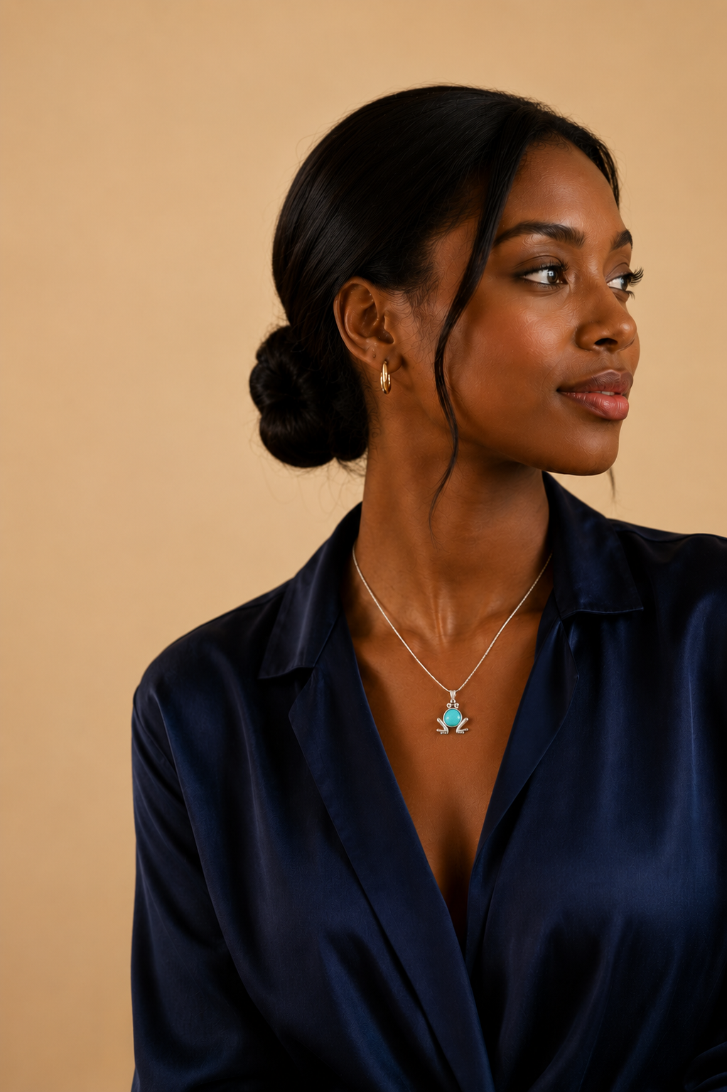 Dark-skinned woman wearing a sterling silver frog necklace with a genuine round amazonite gemstone pendant. Editorial lifestyle portrait featuring an elegant navy satin blouse and warm beige studio background, showcasing a nature-inspired sterling silver necklace for everyday wear