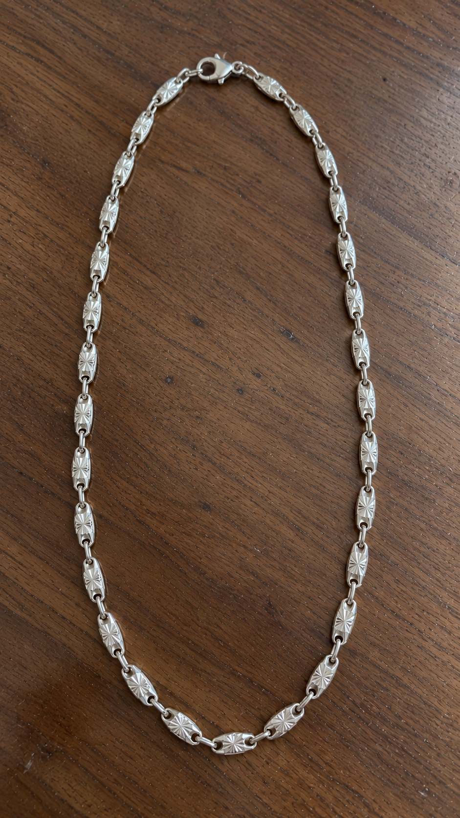Sterling silver starburst chain necklace with engraved links laid on a natural walnut wood table.