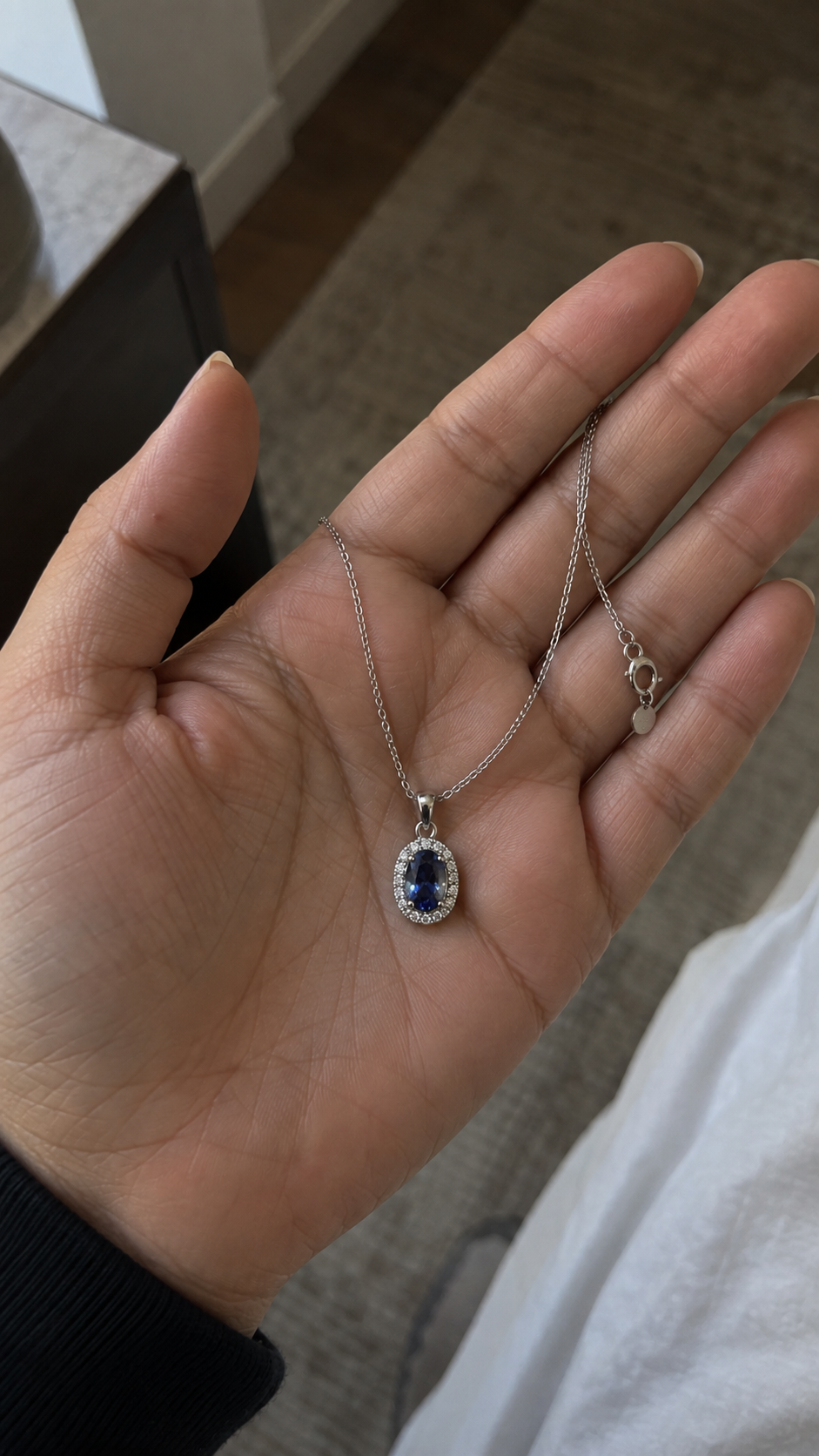 A silver pendant necklace with a deep blue oval gemstone surrounded by small clear stones, held in a person's hand.