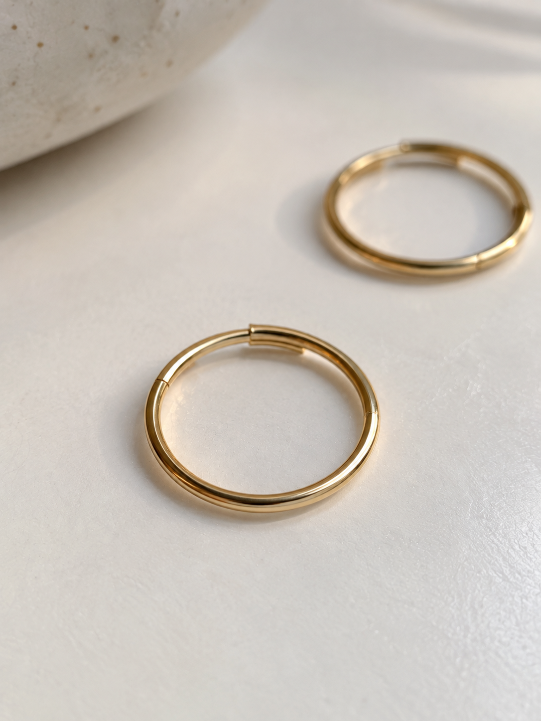 Two thin gold hoop earrings resting on a light off-white surface, with a neutral stone prop partially visible in the top-left corner.